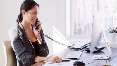 Woman speaking on phone while working at her computer desk, checking data on the PC.